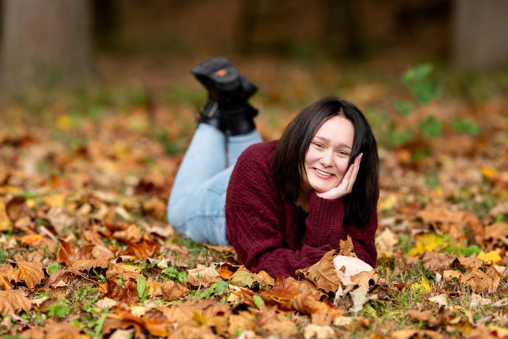 Smiling young woman in maroon sweater lying on autumn leaves in a wooded outdoor setting.