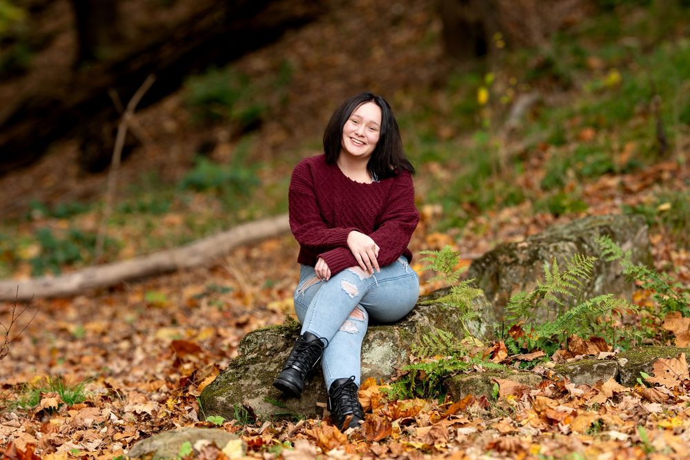 Cheerful young woman in red sweater sitting cross-legged on rock amid autumn leaves in forest.