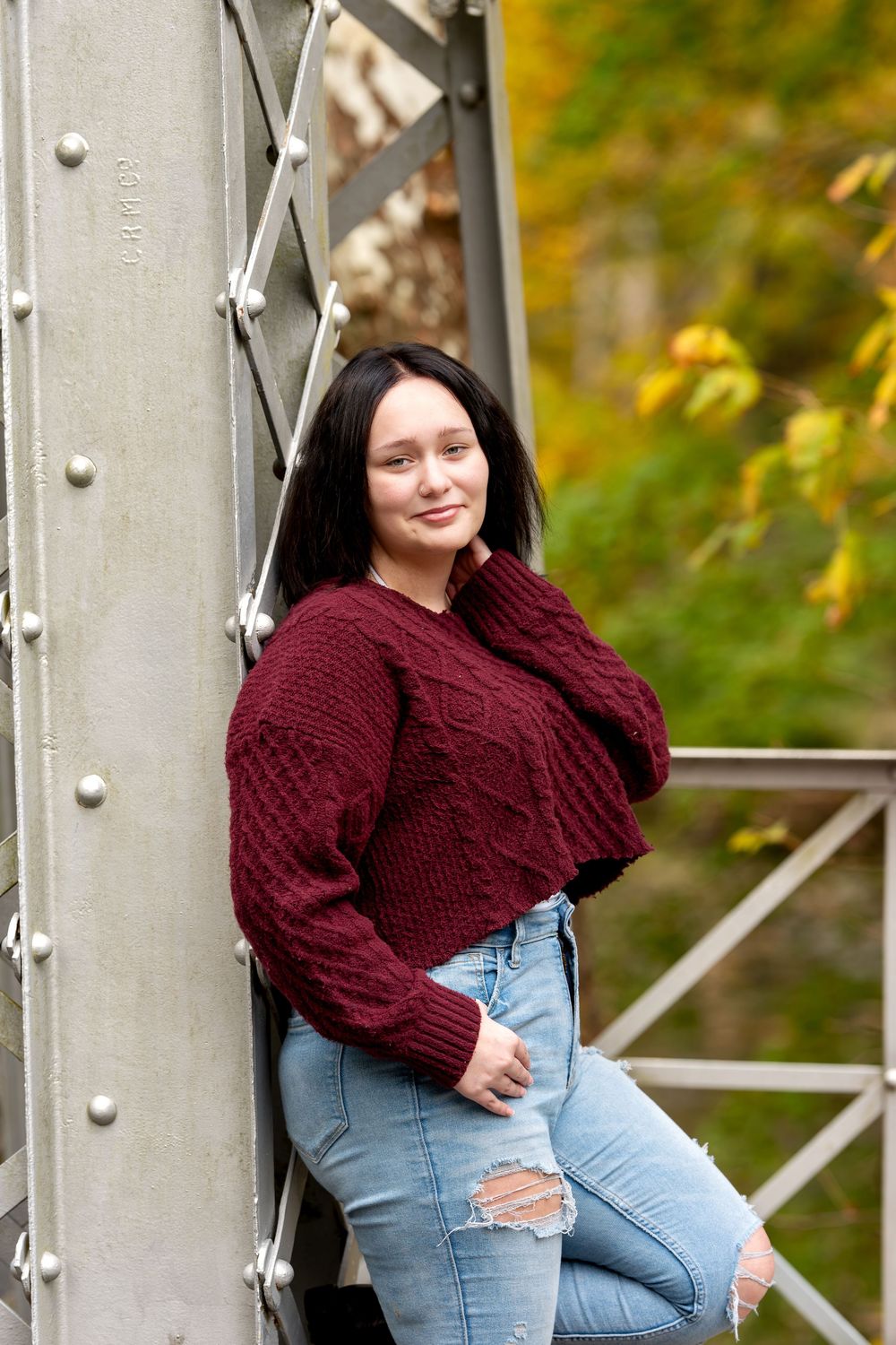Young brunette woman in cable-knit burgundy sweater leaning against metal bridge pillar with fall foliage backdrop.