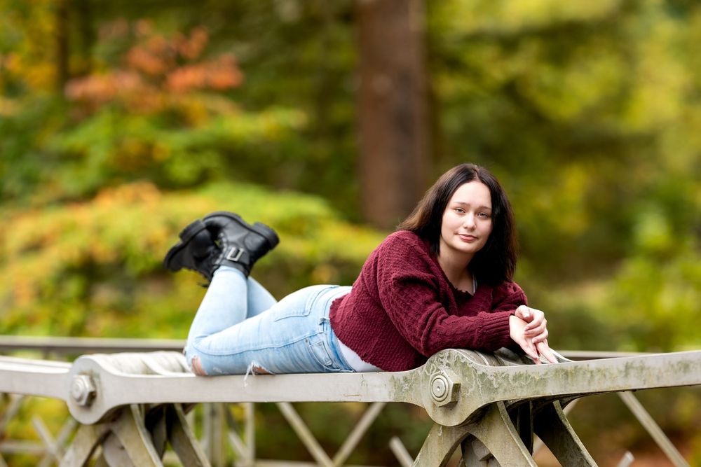 Young brunette woman in burgundy sweater lying on metal bridge railing surrounded by colorful fall foliage.