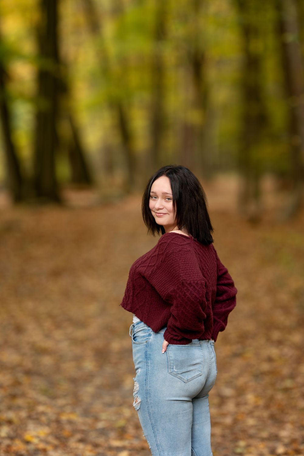 Young woman in maroon sweater and jeans glances back while standing on autumn leaf-covered woodland trail.