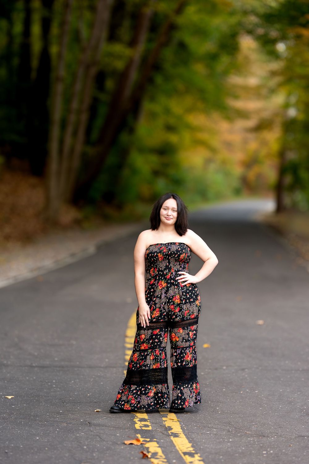 Young woman in a black floral strapless jumpsuit stands confidently on a tree-lined road with fall foliage.