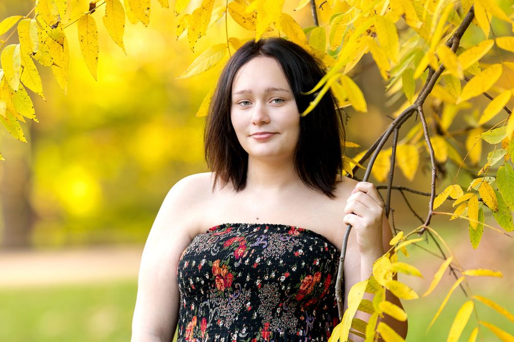 Brunette teen in a strapless floral jumpsuit holds a branch of bright yellow autumn leaves in a sunny park.