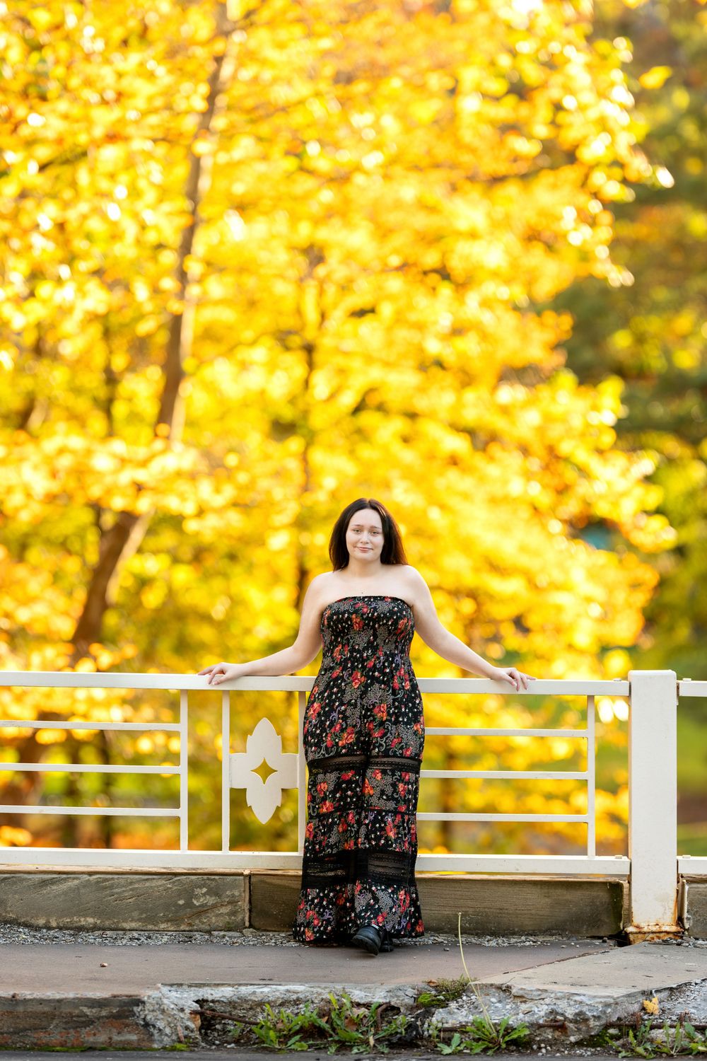 Brunette teen in a strapless black floral maxi jumpsuit stands on a white bridge surrounded by golden fall trees.