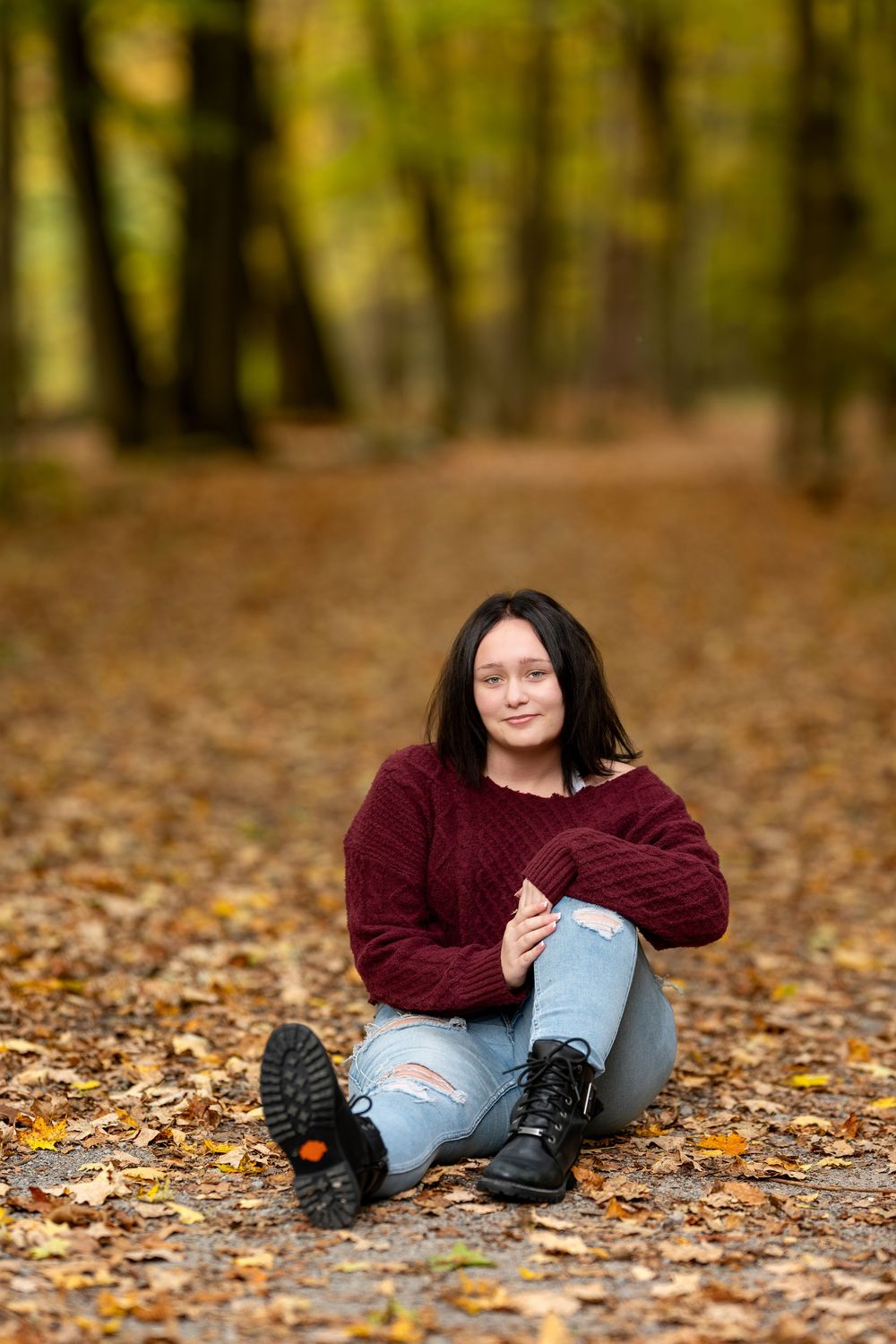 Young woman in red sweater and distressed jeans sits cross-legged on autumn leaf-covered forest path.