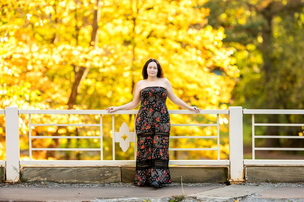 Young woman in black floral maxi dress standing on white bridge with vibrant yellow fall trees behind her.