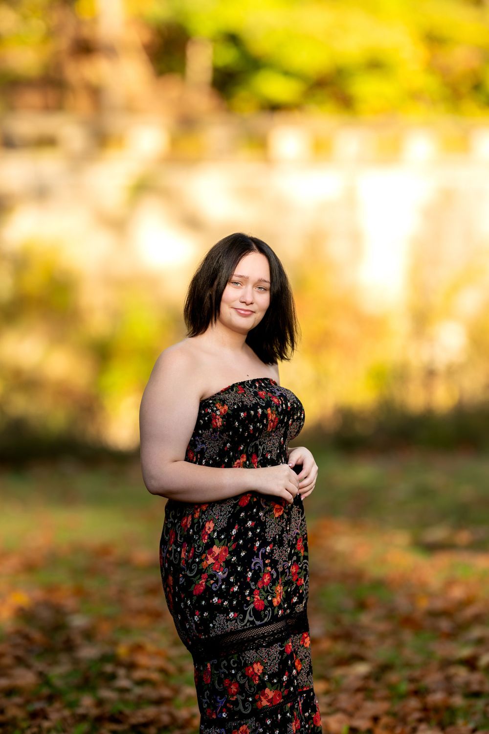 Woman in black floral strapless dress standing outdoors holding hands together in warm autumn light.