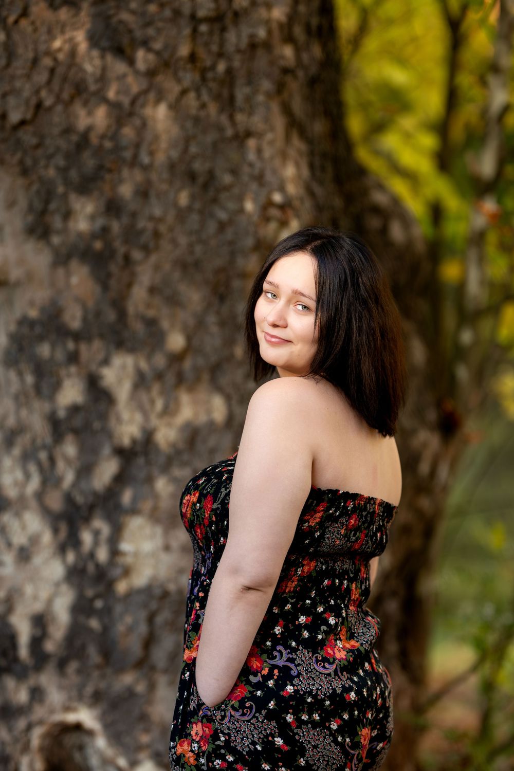 Woman in black floral strapless dress standing beside large tree trunk glancing over her shoulder.