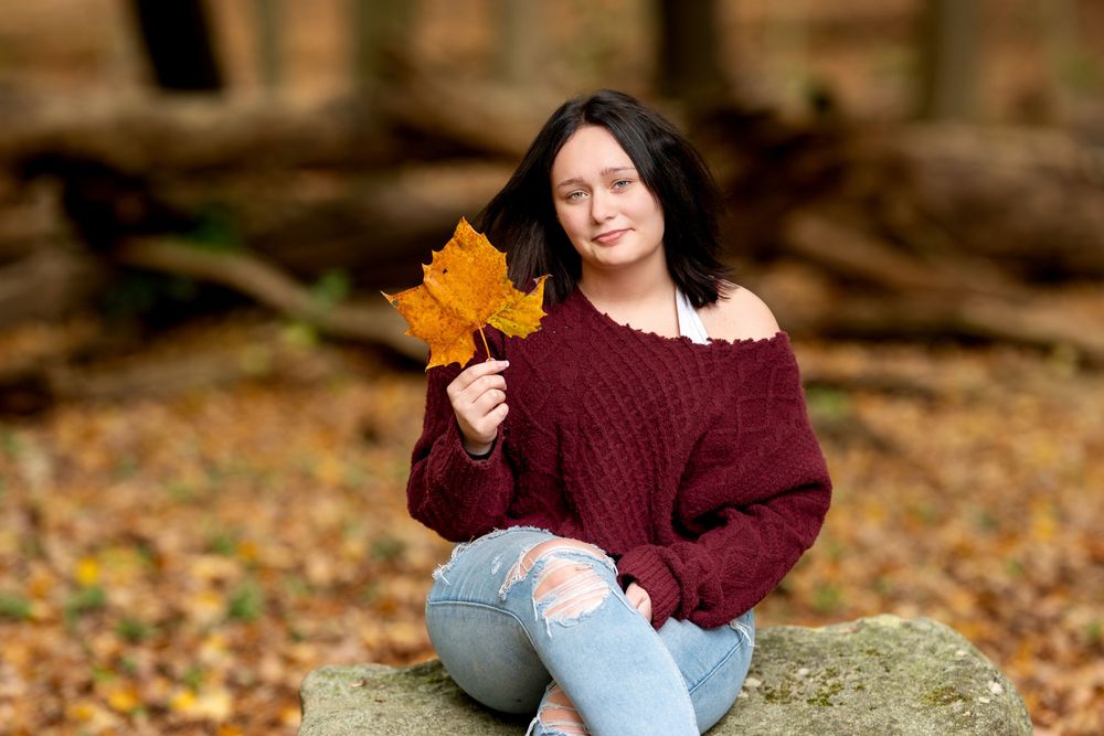 Brunette woman in burgundy off-shoulder sweater sits on rock holding golden maple leaf in autumn forest.