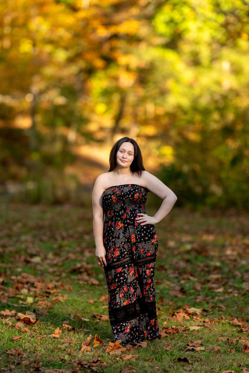 Young brunette woman in black floral maxi dress posing in colorful autumn forest.