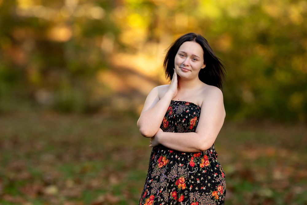 Young woman in floral strapless dress poses outdoors in autumn park with colorful fall foliage background.