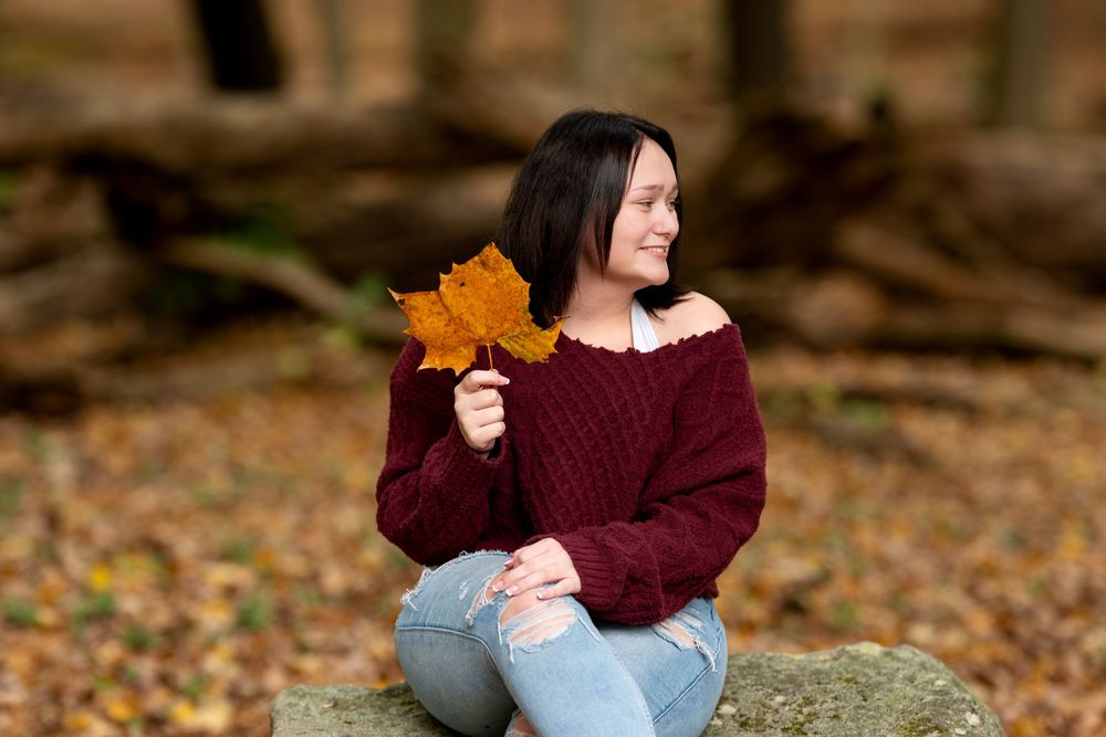 Smiling woman in burgundy off-shoulder sweater holds autumn maple leaf while sitting in fall forest setting.