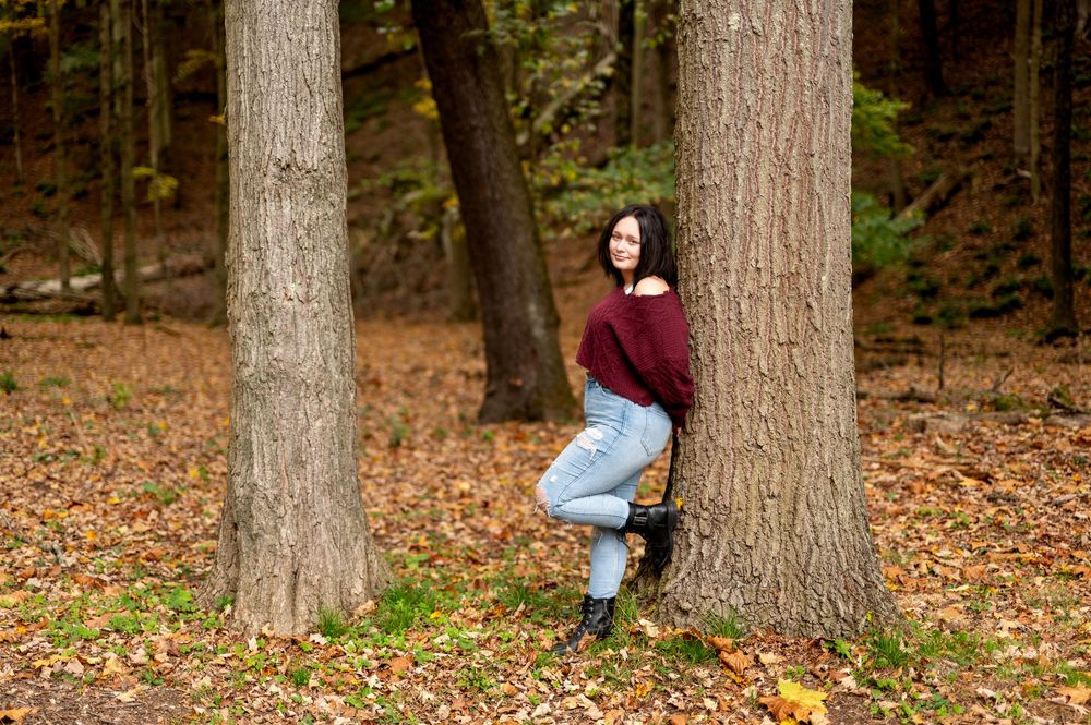 Dark-haired teen in burgundy sweater and ripped jeans standing between trees on autumn leaf-covered ground.