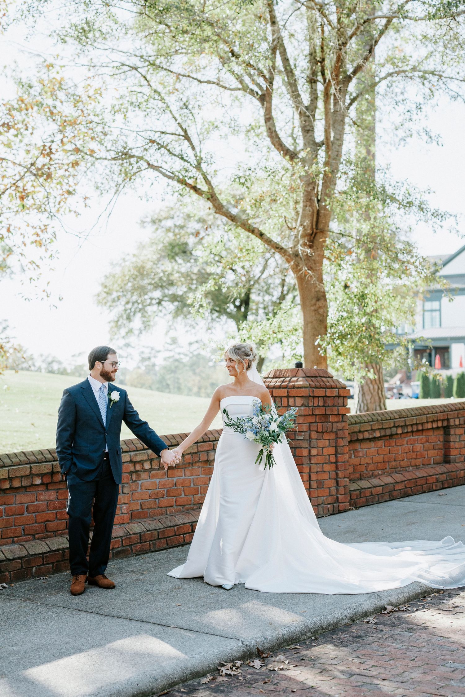 A couple in wedding attire hold hands while standing by a brick wall under a large tree on their special day.