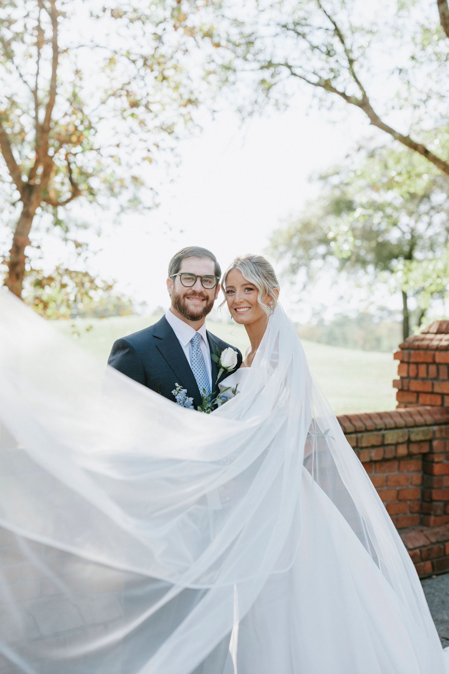A flowing white wedding veil catches the breeze as a couple shares a romantic moment in front of a brick wall.
