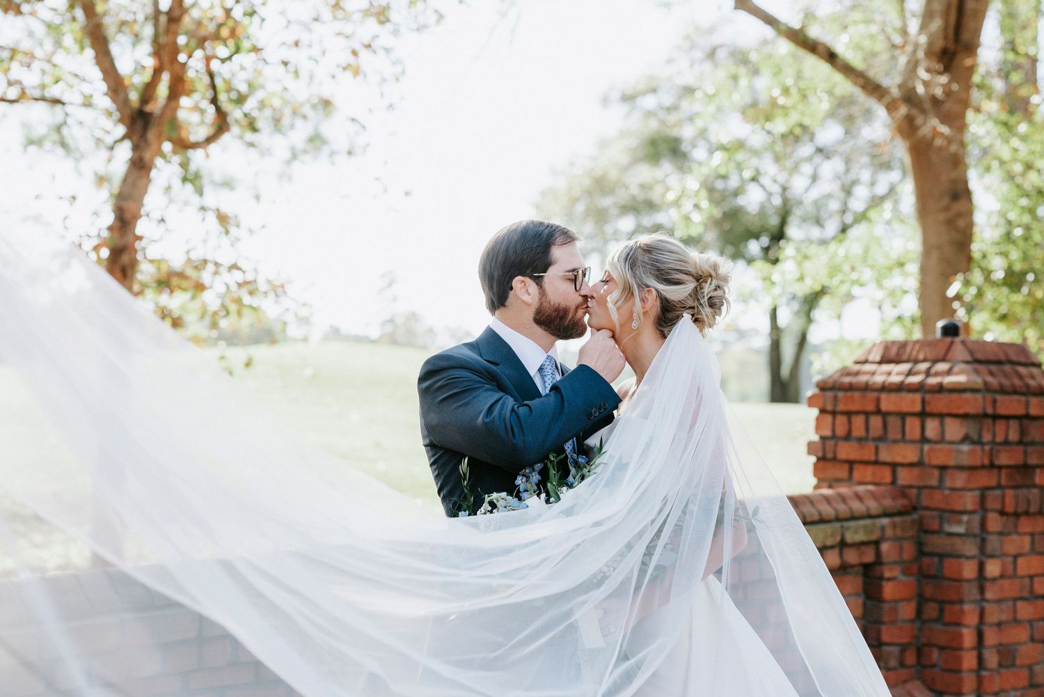 A bride in a flowing white veil stands in a garden setting as the wind catches her dress and veil.