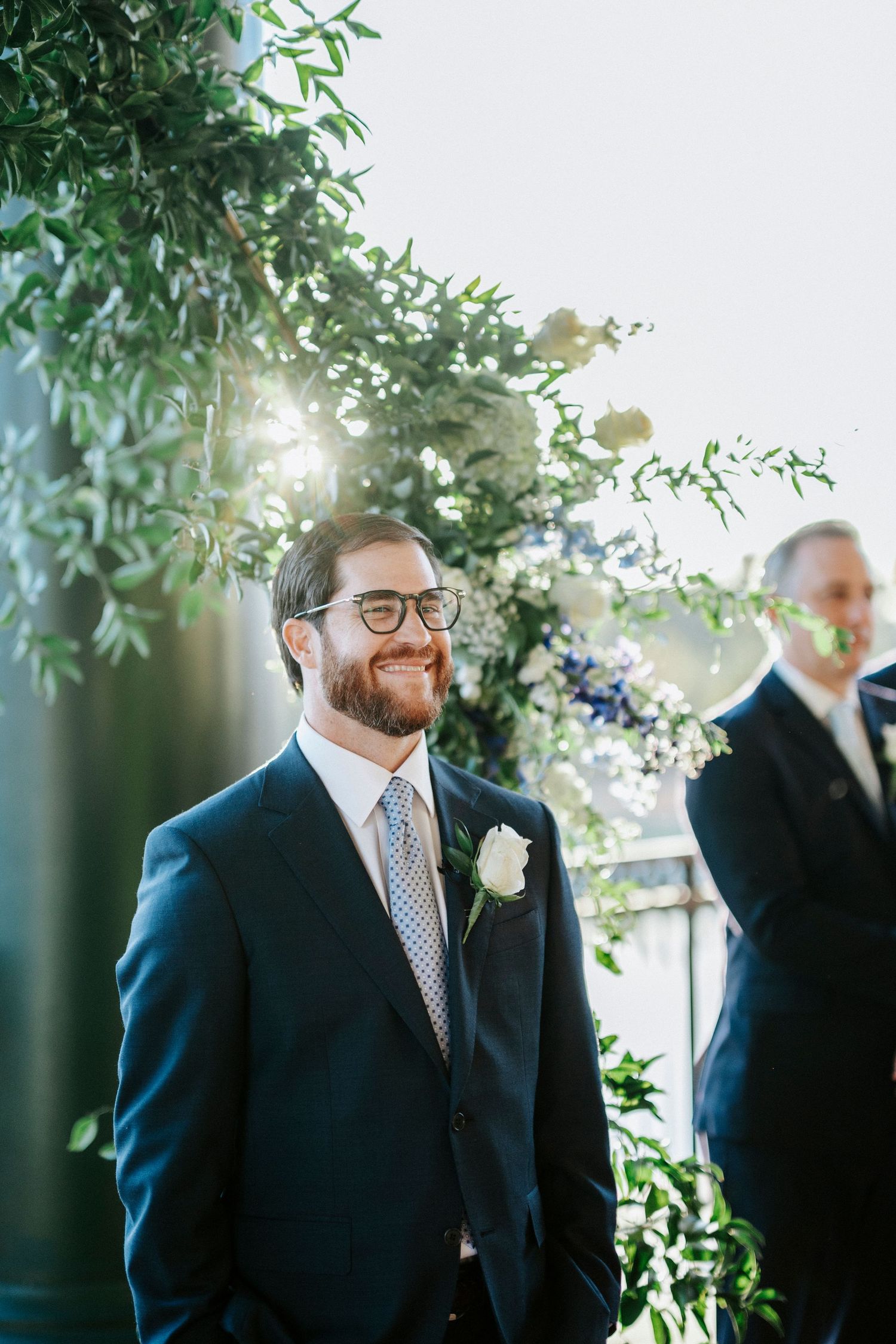 A groomsman stands at an outdoor wedding ceremony with sunlight filtering through green foliage and white floral decor.