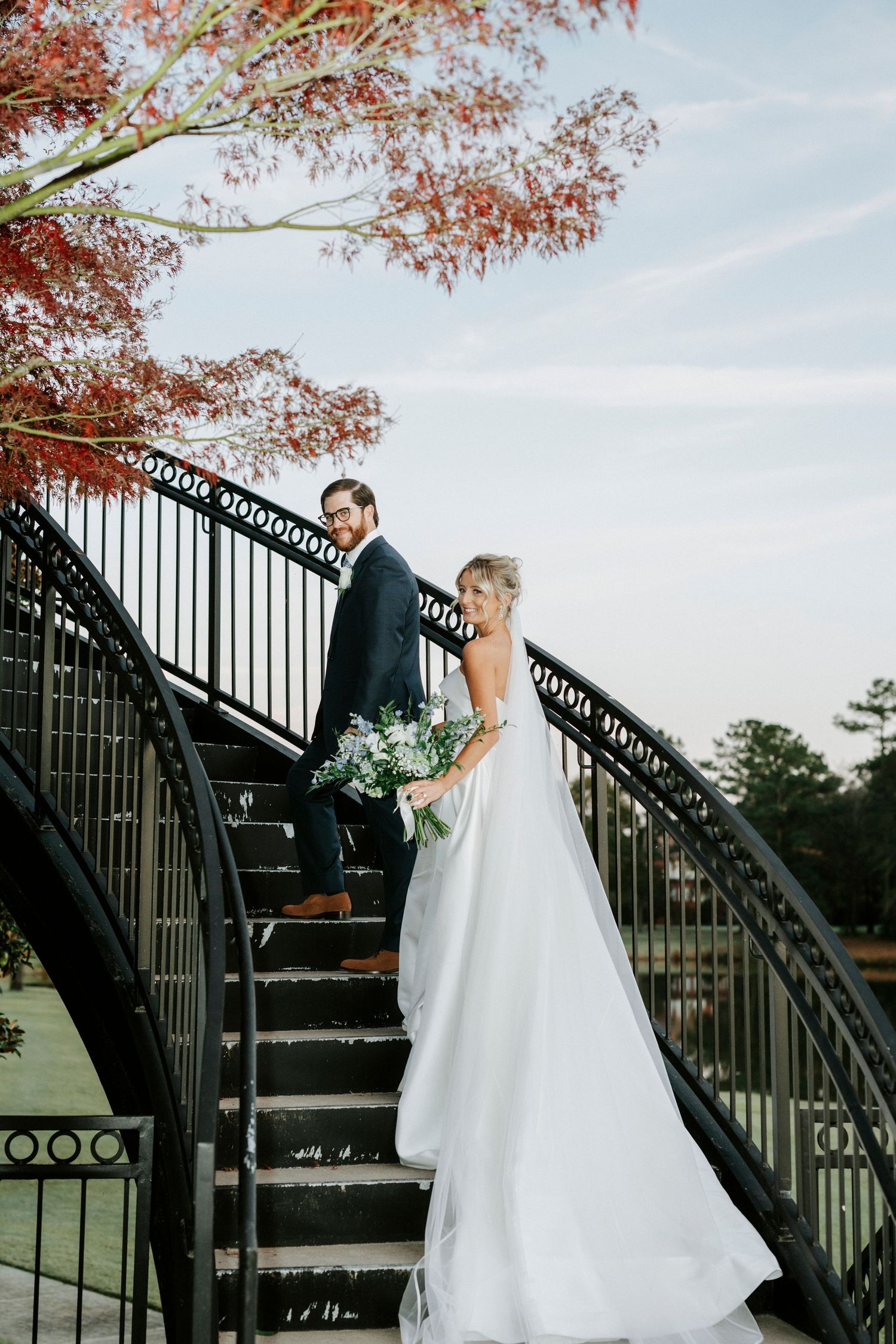 A romantic wedding couple poses on a grand staircase with autumn foliage and decorative iron railings in the background.