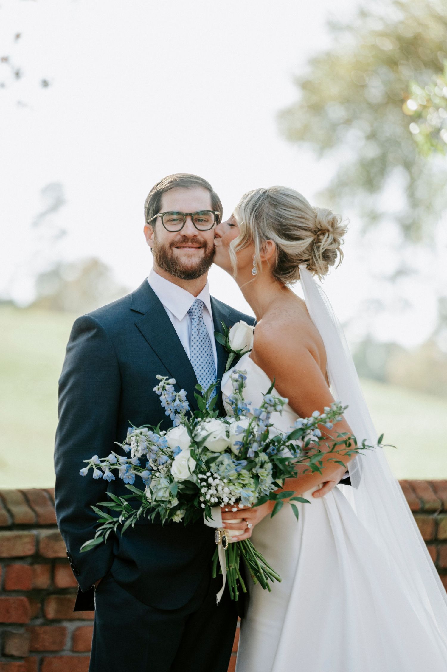 A romantic wedding couple shares tender moments with white floral bouquet against a brick wall backdrop.
