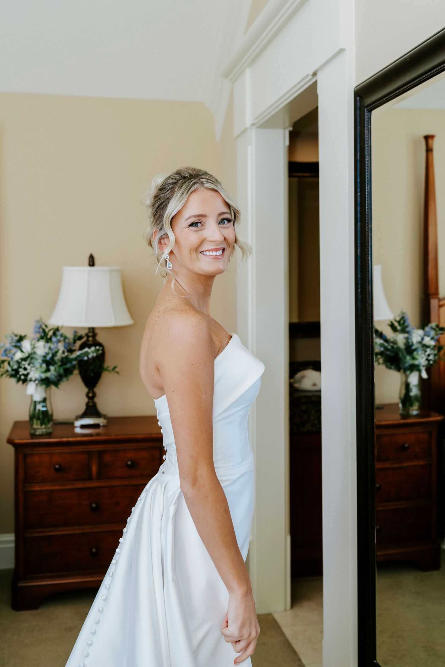A bride in a classic white strapless wedding gown smiles while standing in a bedroom with traditional wooden furniture.