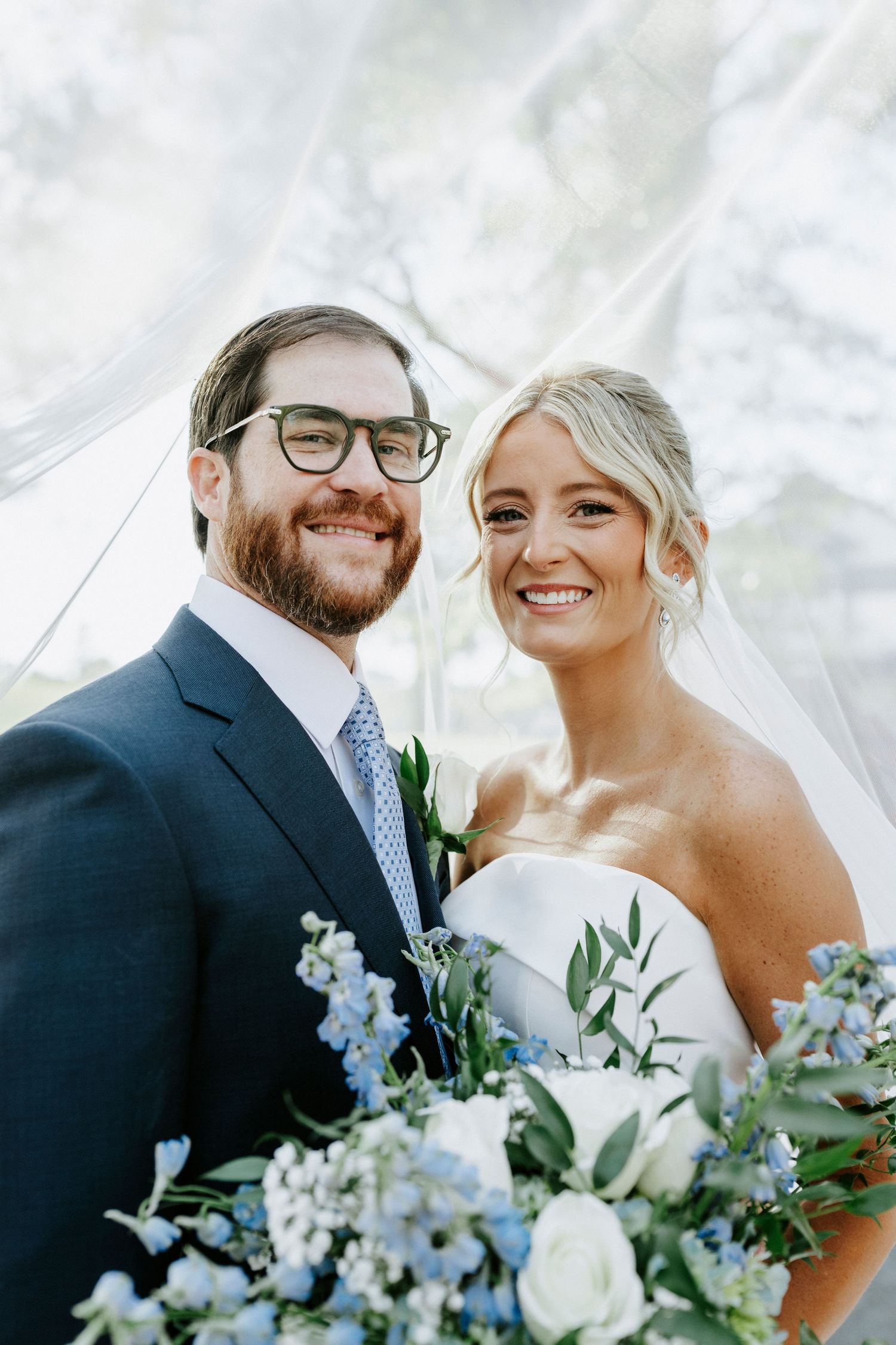 Wedding couple poses with delicate blue and white floral bouquet against bright background.