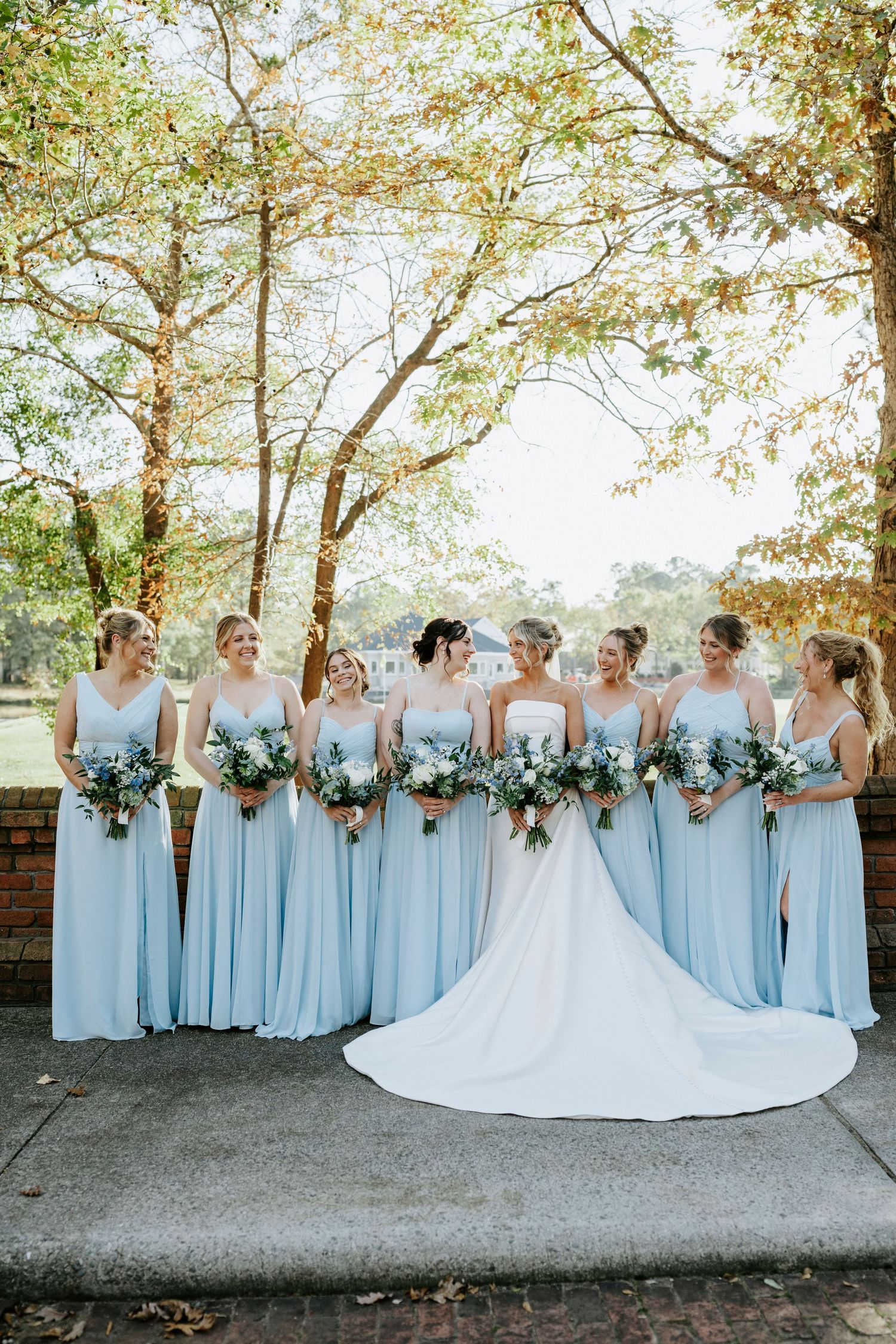 A wedding party in light blue bridesmaid dresses and a white bridal gown pose together against a rustic outdoor wall.