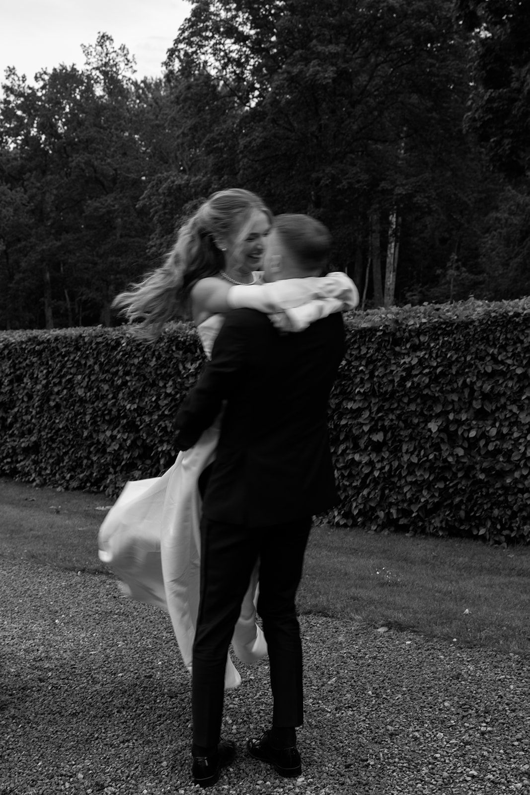 A couple dancing together in an intimate embrace on a garden path with a hedge wall in black and white.
