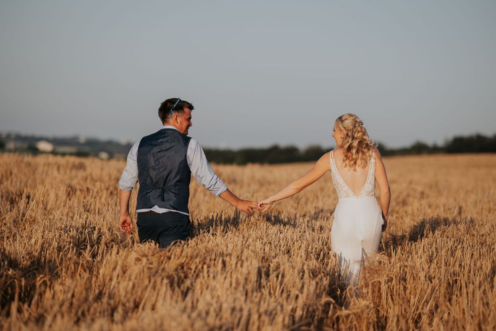 Bride and groom holding hands walking through a golden wheat field during sunset on their wedding day.