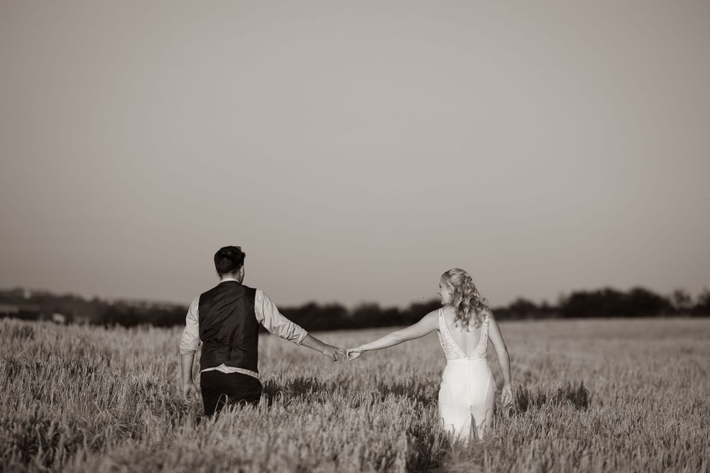 Black and white photo of bride and groom holding hands walking through an open field at dusk.