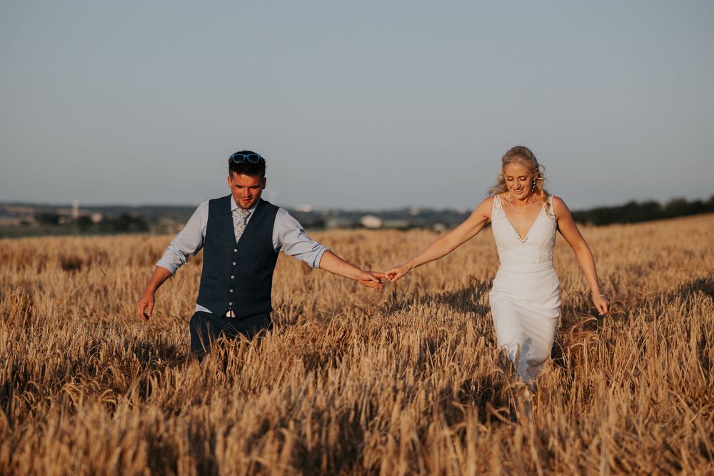 Bride and groom laughing and walking hand in hand through a sun-drenched golden wheat field.
