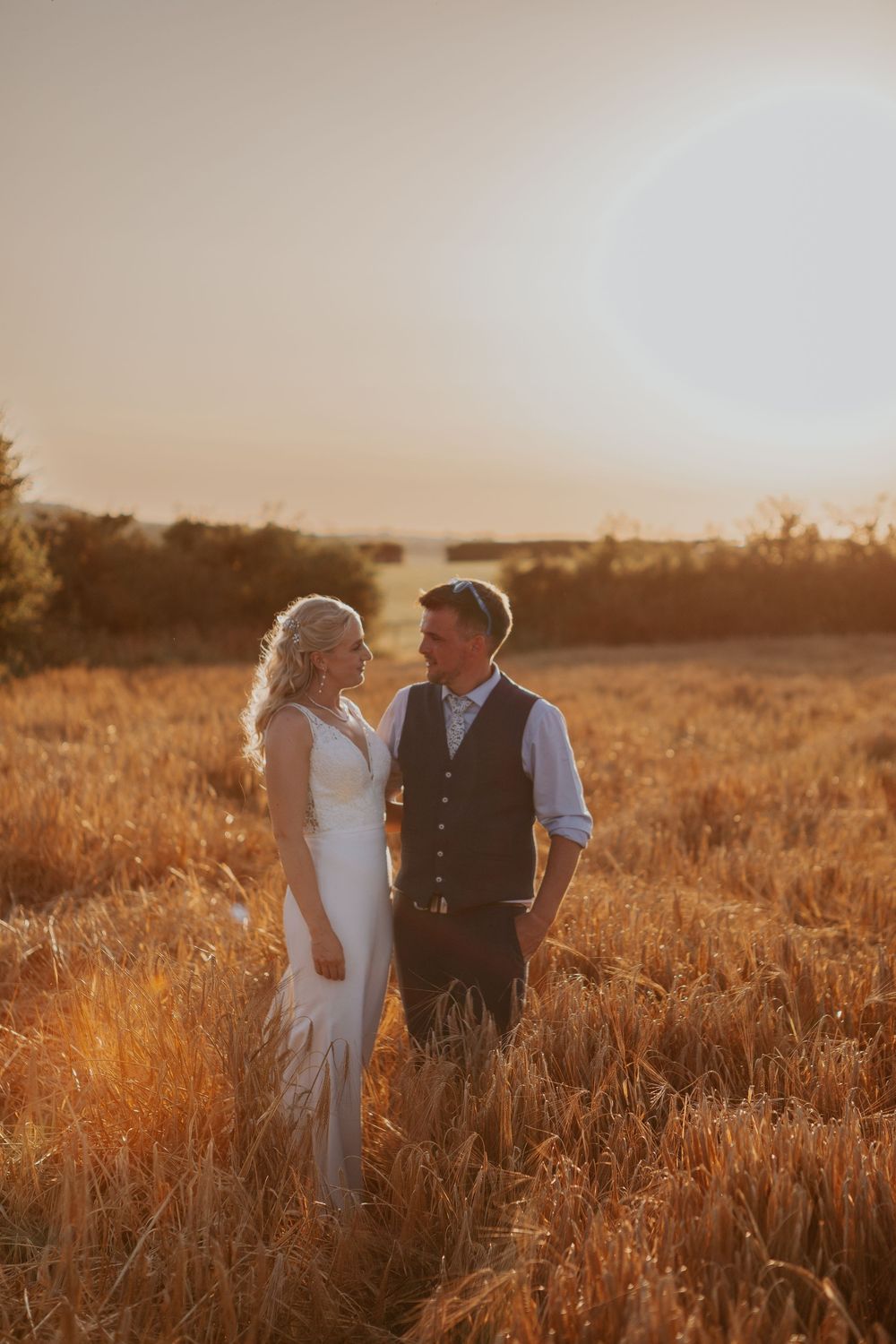 Bride and groom gazing at each other lovingly while standing in a glowing golden wheat field at sunset.