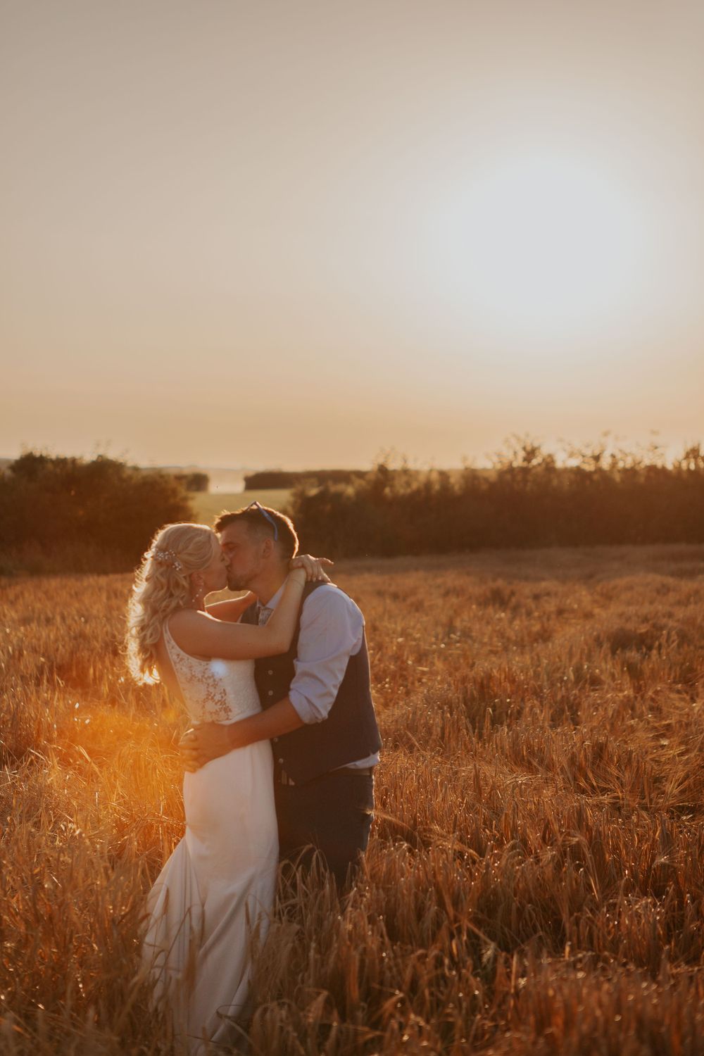 Bride and groom sharing a golden hour kiss in a wheat field bathed in warm sunset light.