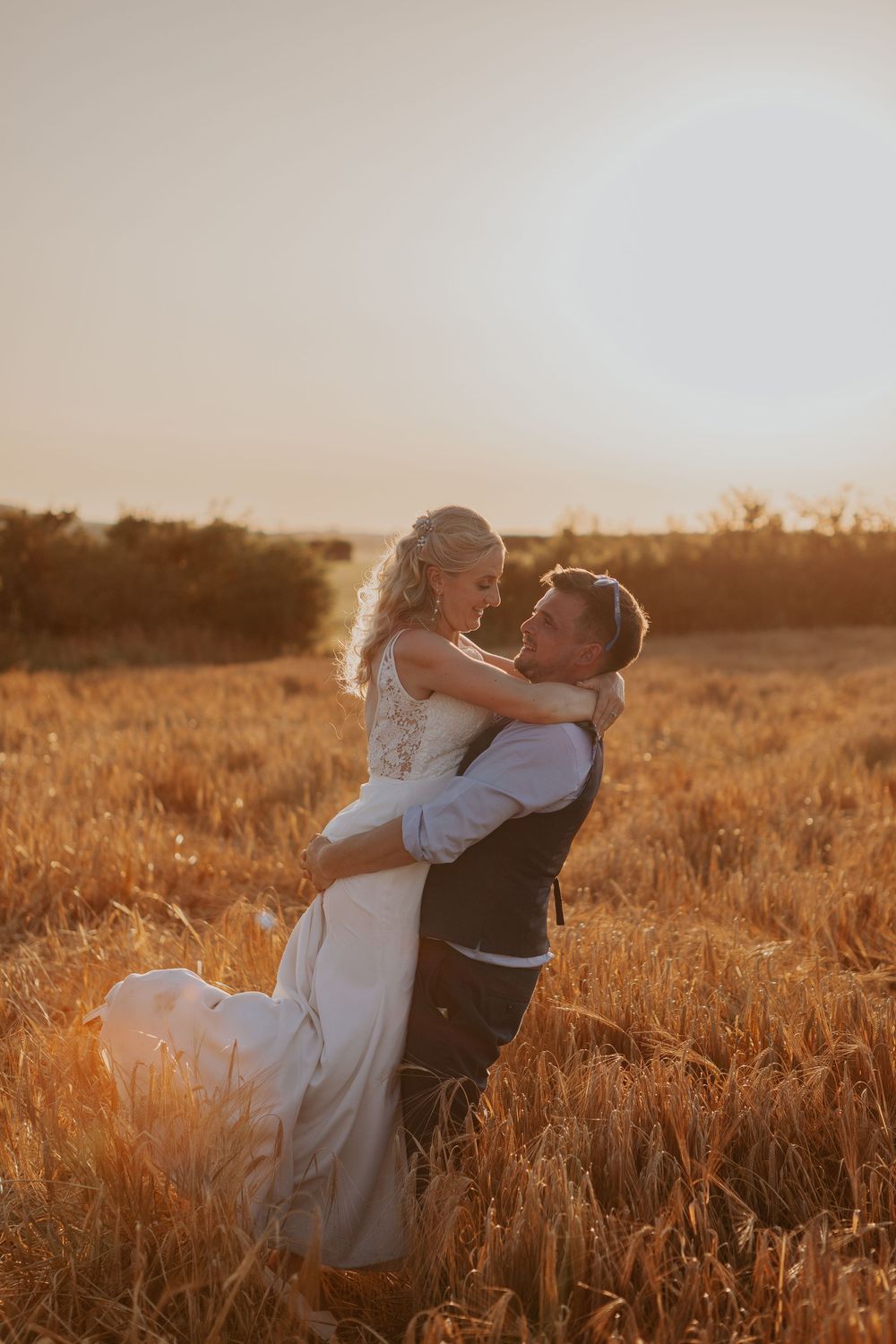Groom lifting his smiling bride in a warm golden wheat field during a breathtaking sunset.