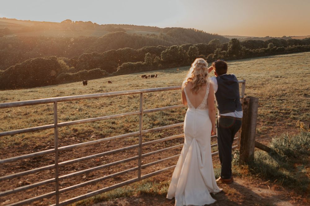 Bride and groom standing at a farm gate enjoying a scenic countryside view at golden hour.