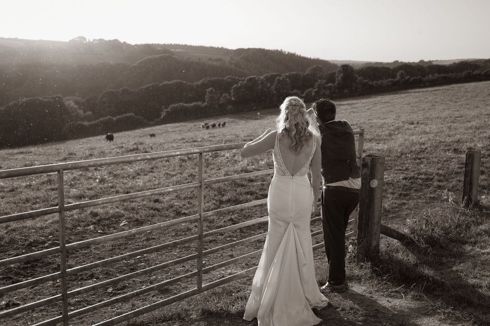 Black and white photo of bride and groom standing at a farm gate overlooking rolling countryside hills.