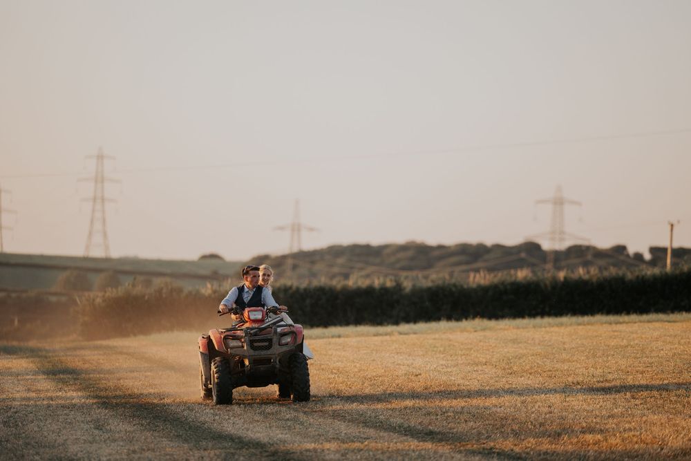 Bride and groom riding a red quad bike through a dusty farm field at golden hour sunset.