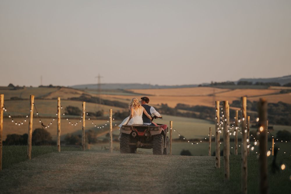 Bride and groom seated on a quad bike surrounded by fairy lights in a scenic countryside vineyard.
