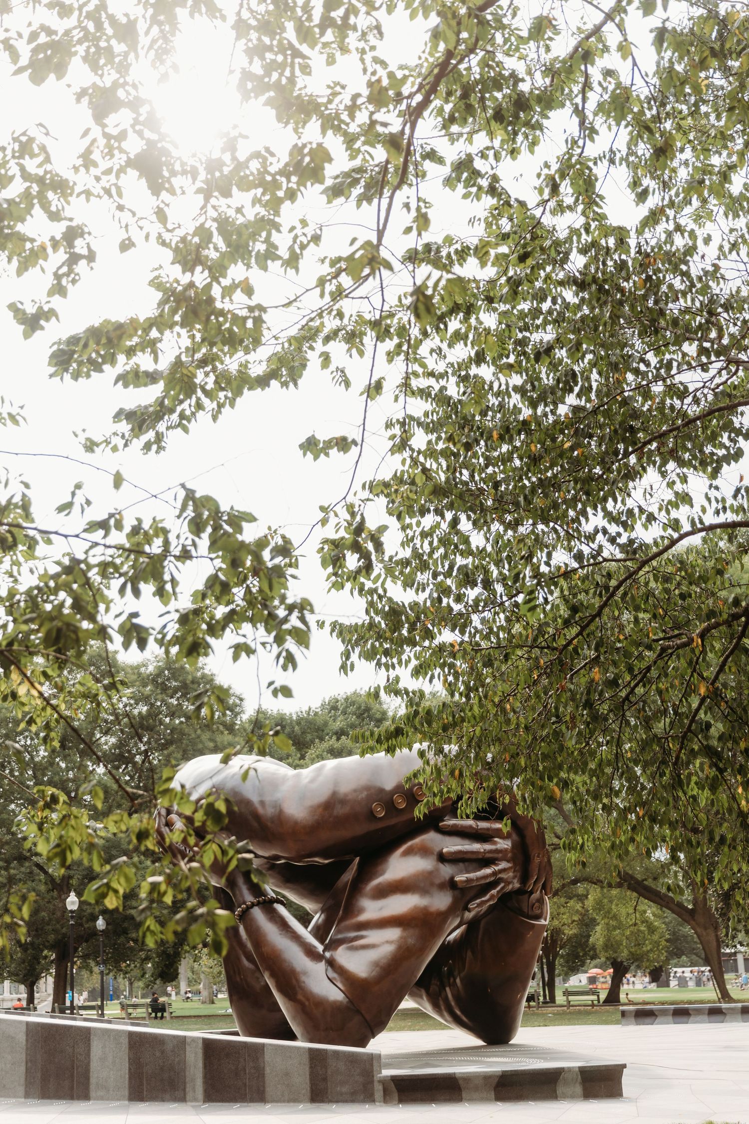 Large bronze sculpture of reclining abstract figures in a park setting with trees and benches.