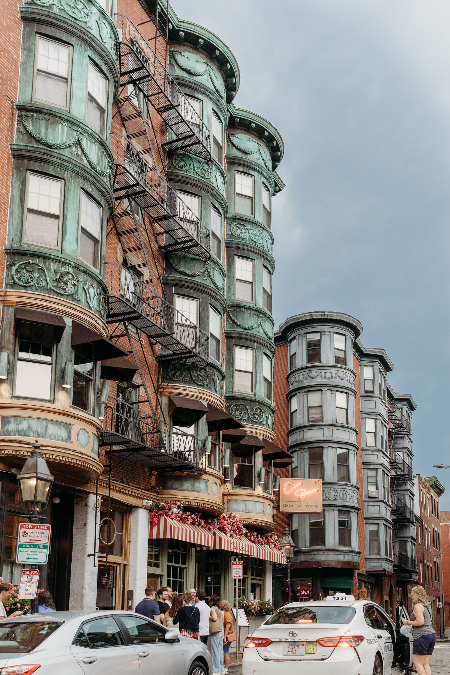 Historic green-trimmed Victorian buildings line a street in Boston's North End neighborhood with parked cars below.