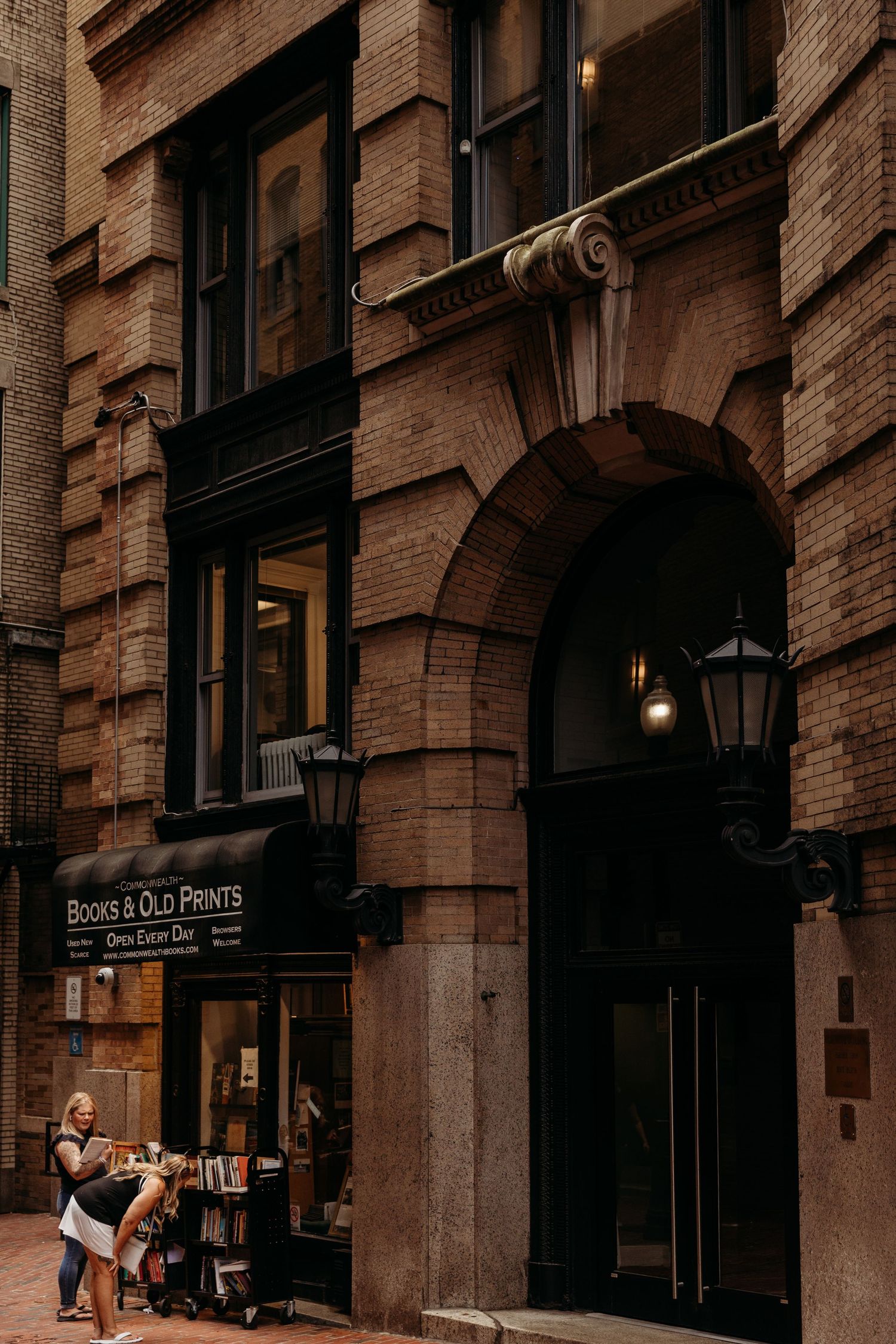 Historic brick building exterior with arched entrance and storefront sign for Books & Old Prints shop on a moody city street.