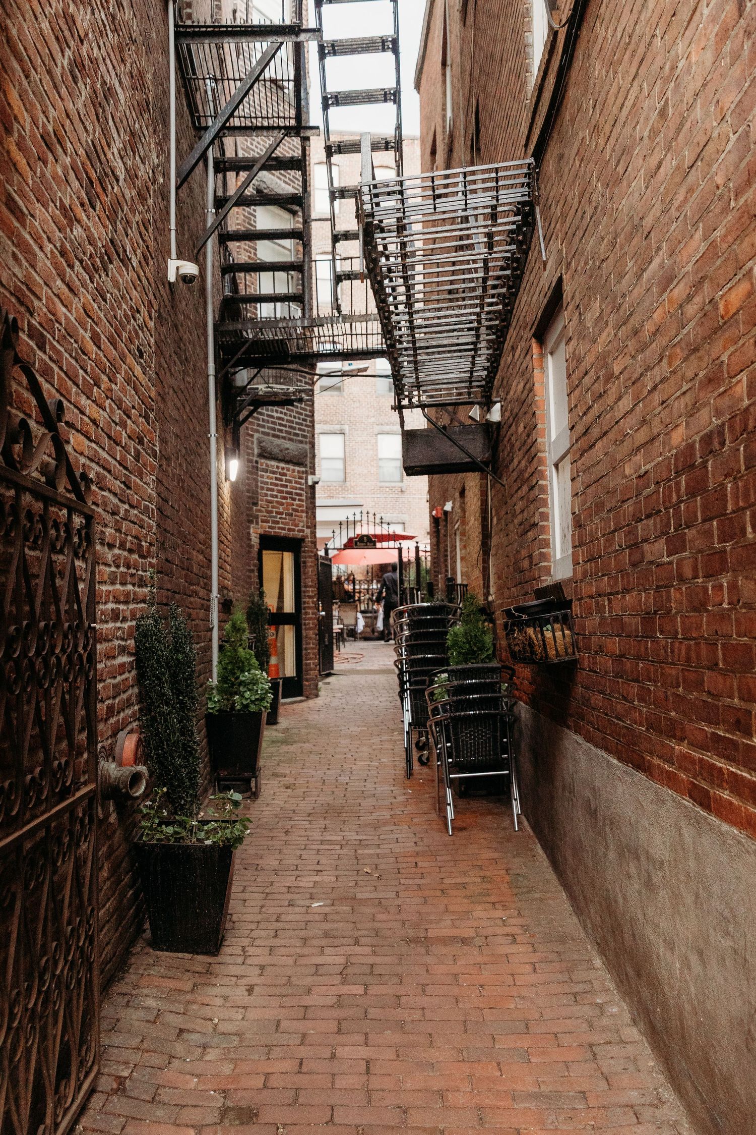 Narrow brick alley with fire escapes and metal railings leads down a historic urban passageway.