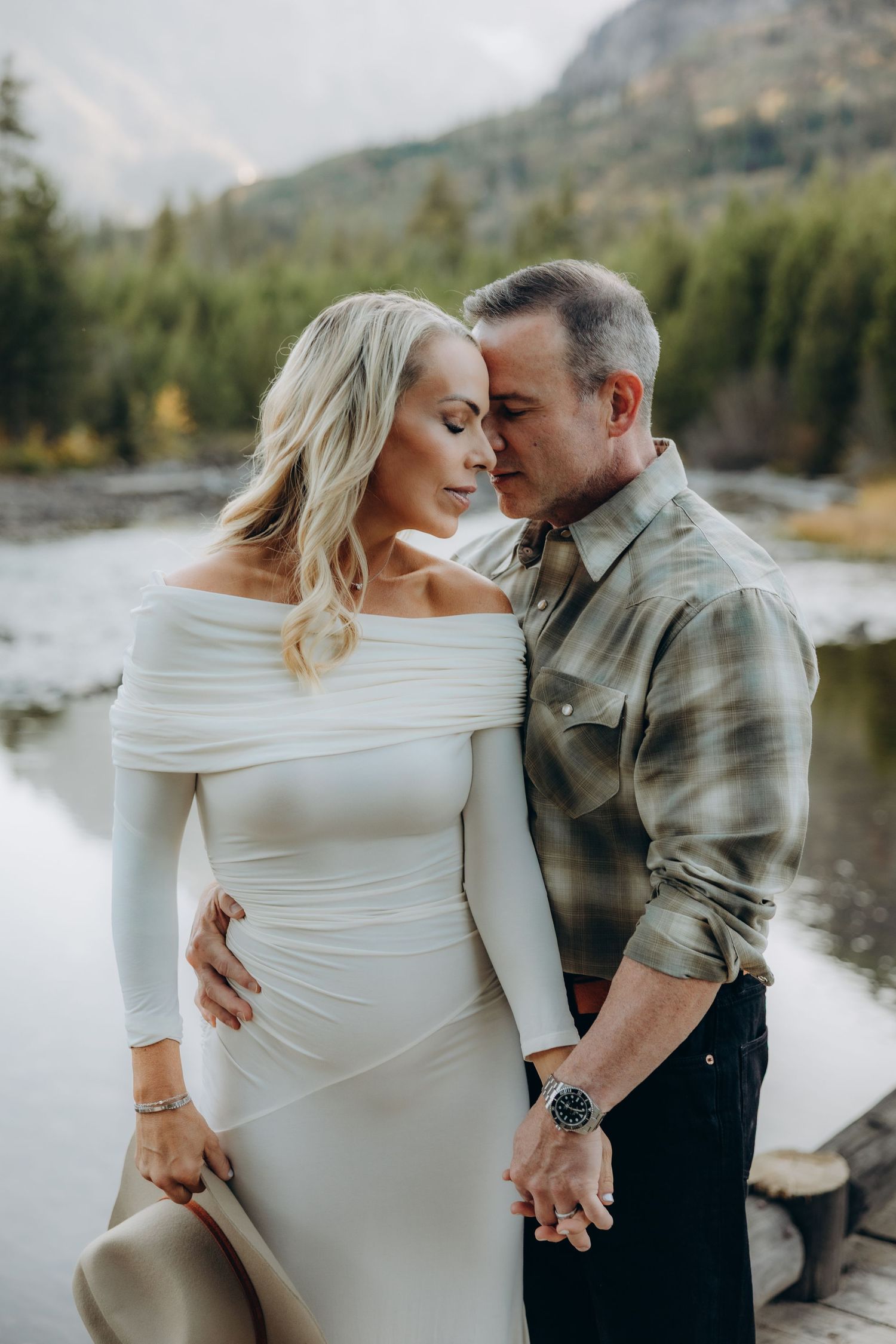 Intimate maternity photoshoot by a lake with couple in white dress and casual attire sharing tender moments.