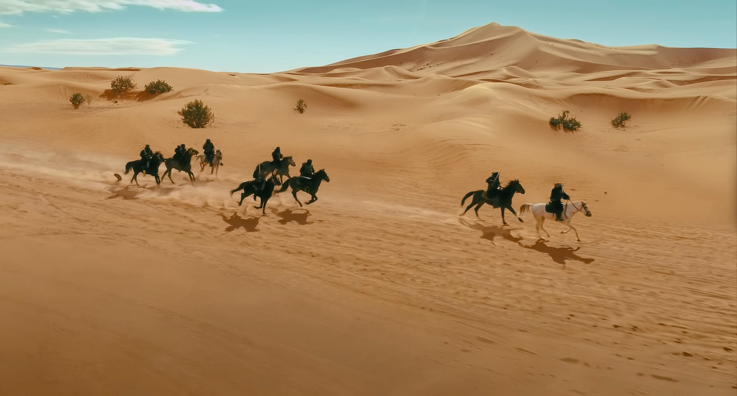 Silhouettes of camel riders traverse golden sand dunes in the Sahara Desert under a bright blue sky.