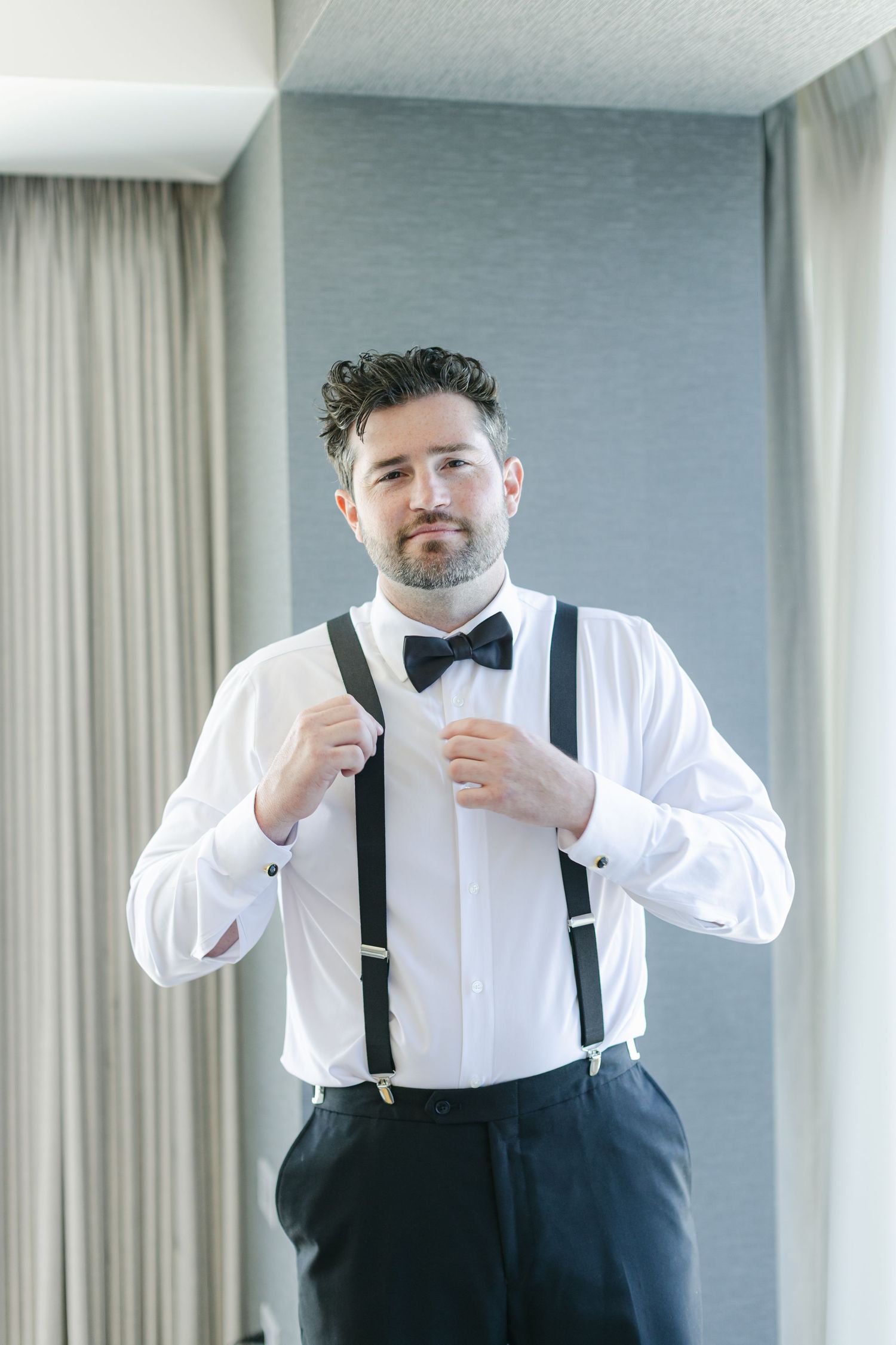 A person in a formal white shirt and black suspenders adjusts their tie while getting dressed in a hotel room.