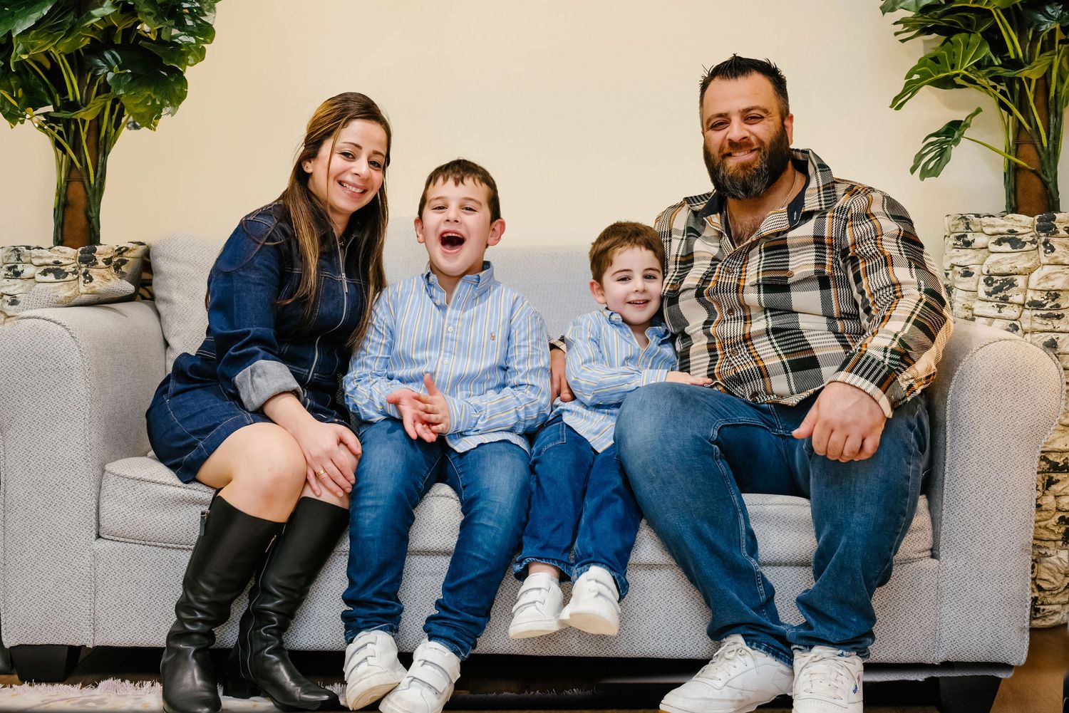 A family of four sits together on a gray couch, wearing casual denim and plaid clothing in an indoor setting.