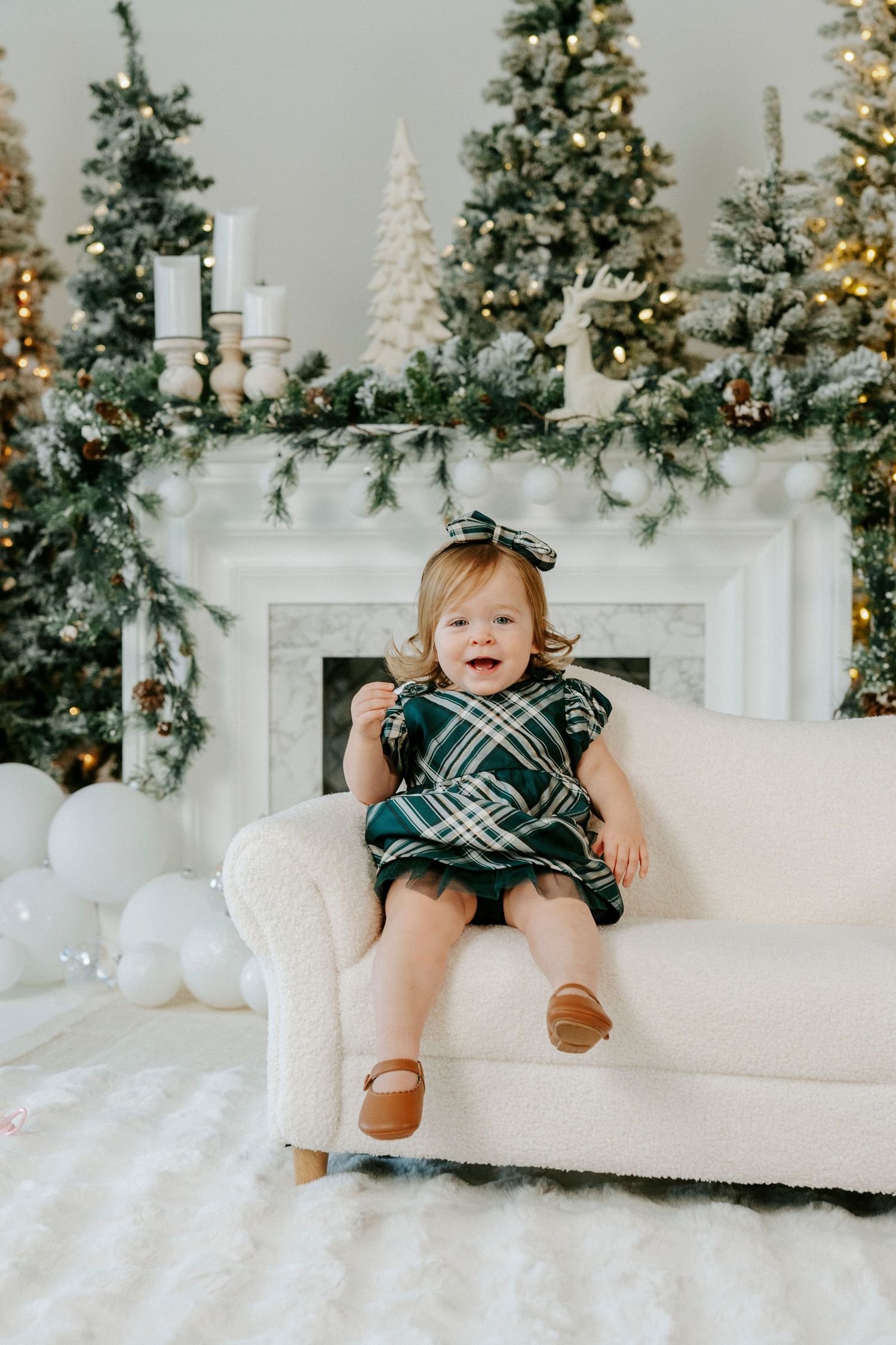 Festive holiday scene with decorated Christmas trees and garland on white mantle while small child sits on cream armchair.