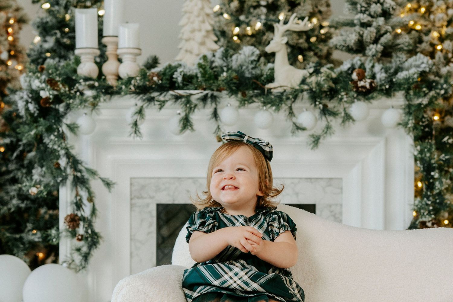 A child in a plaid holiday dress sits on a white couch in front of a decorated Christmas fireplace mantel.