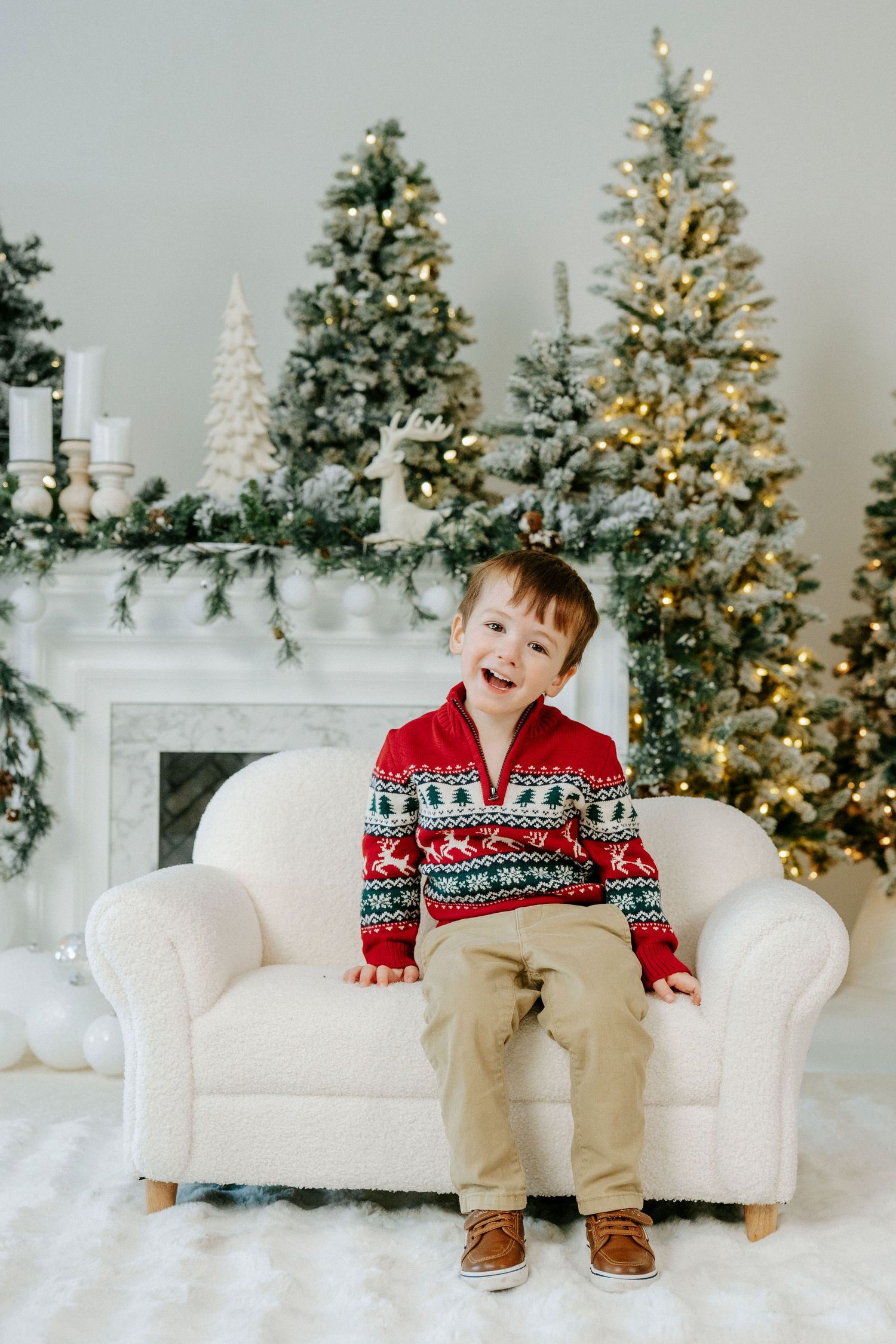 A child in a festive red sweater sits on a white couch surrounded by decorated Christmas trees and holiday decor.
