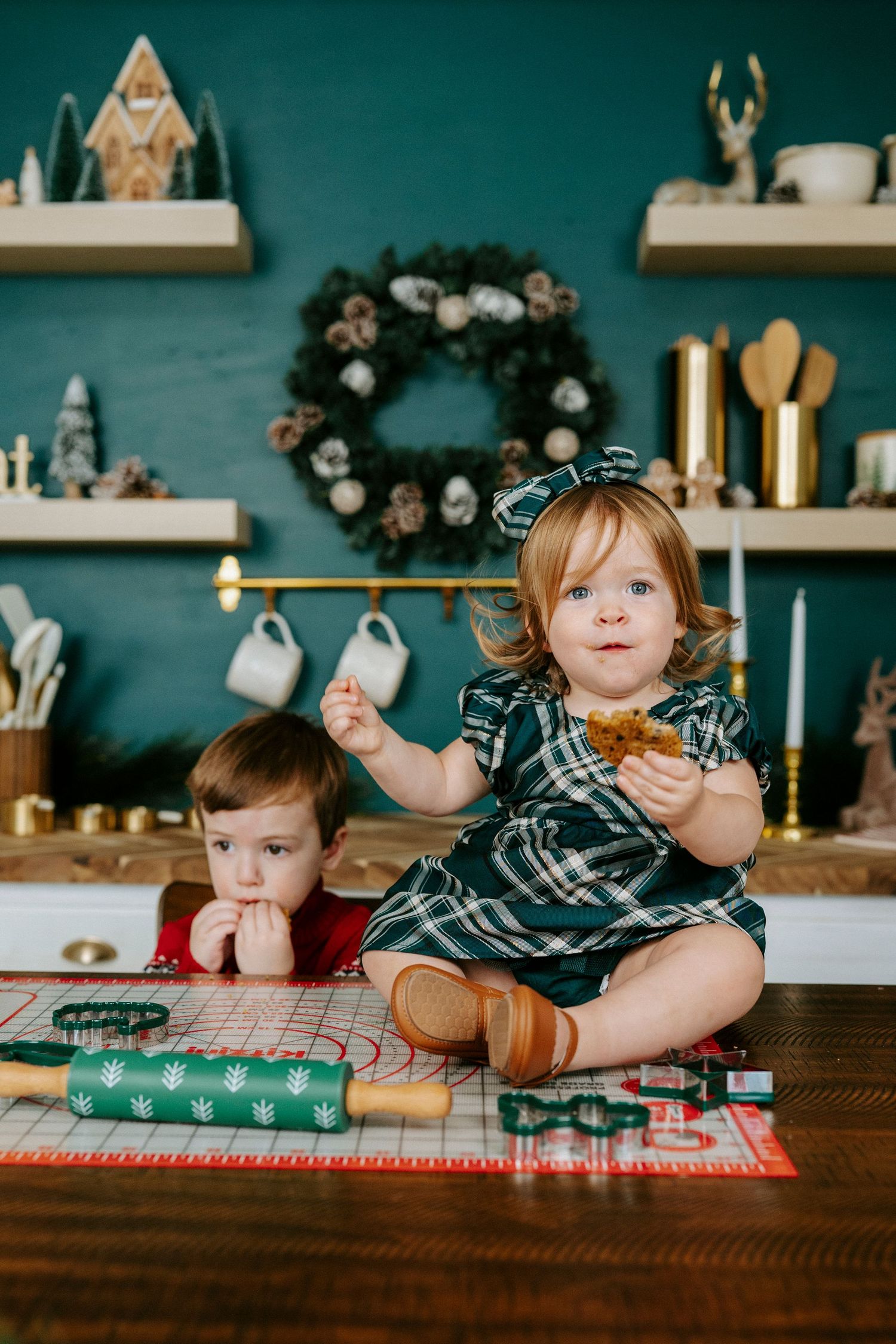 Two children sit on a wooden counter doing holiday baking with cookie cutters and festive decor on teal walls.