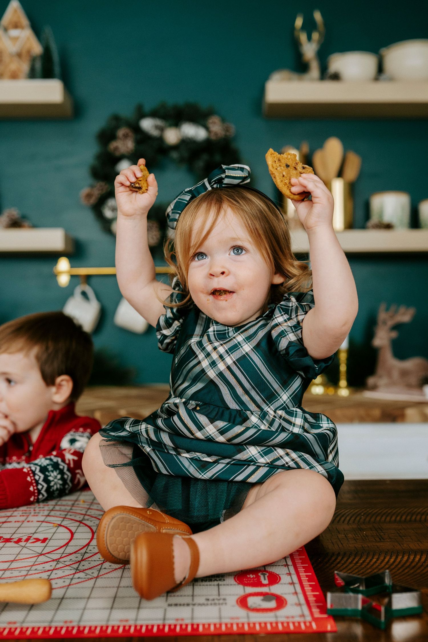 Young children play and interact on a wooden table with a red and white checkered tablecloth in a cozy kitchen setting.